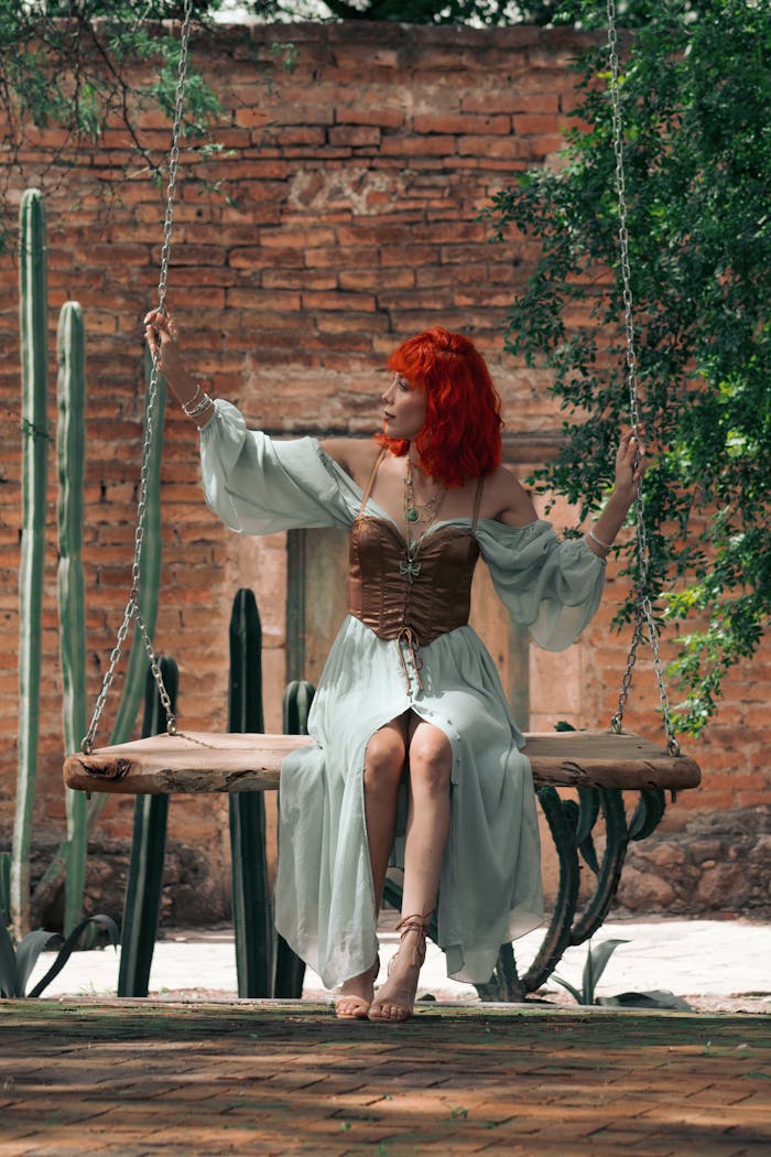 A redhead woman in a vintage dress sits on a swing outdoors amidst tall cacti.